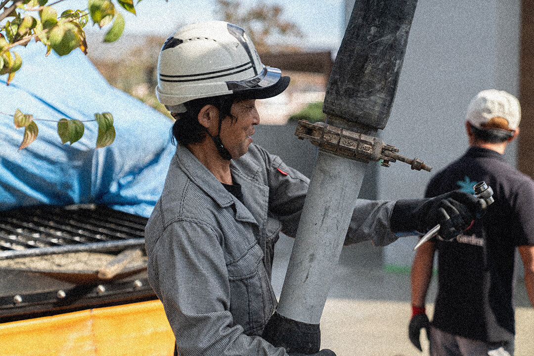 作業服を着てヘルメットをかぶった男性が、コンクリート圧送用の太い配管を支えるように持ちながら、横を向いて作業している。
